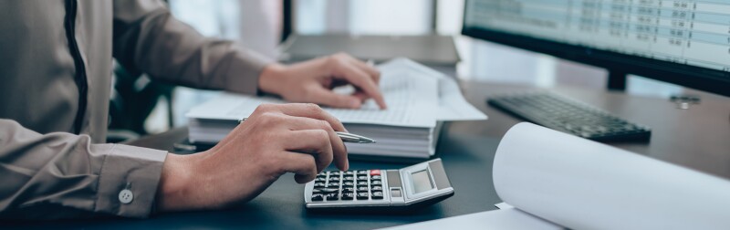 Auditor working at a desk with a stack of spreadsheets and a calculator.