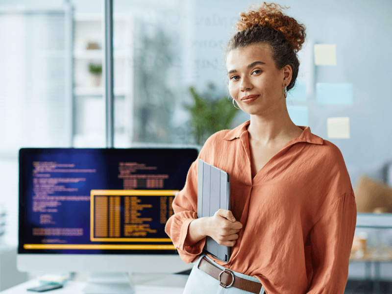 Woman in orange shirt holding tablet standing in front of desktop monitor