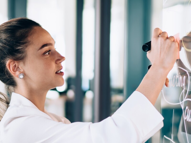 Woman writes with marker on glass board while man looks on.