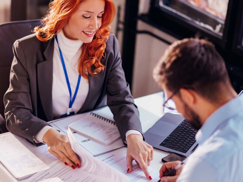 A female investment banker sits at a desk advising a male client where to sign on a document.