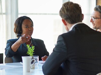 Five diverse professionals are sitting around a table in a naturally lit conference room. They are having a discussion.