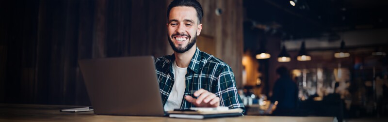 Smiling man in casual attire working on a laptop in a warmly lit cafe