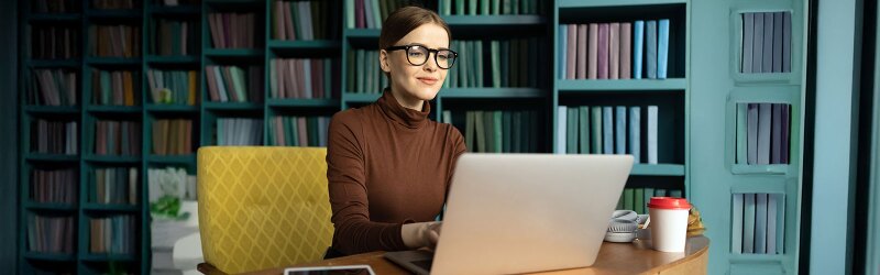 A woman in glasses studying on a laptop with bookshelves in the background, representing an online education program for teaching.
