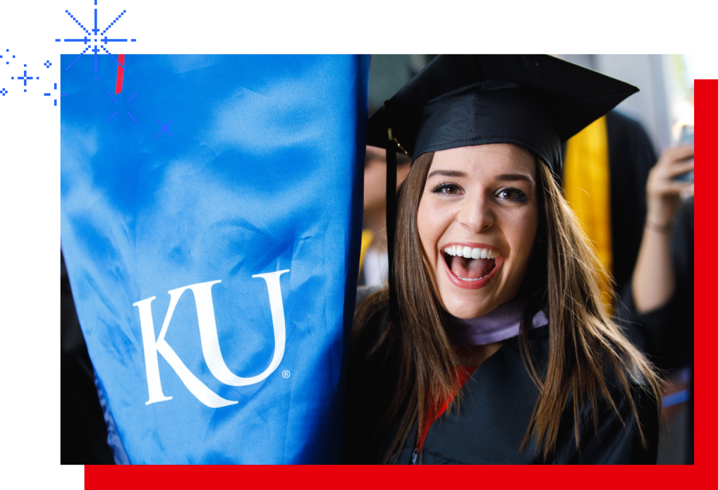 Smiling graduate in cap and gown holding a KU banner during a graduation ceremony.