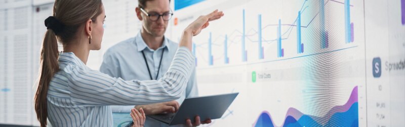 Caucasian male and female data scientists with laptop and tablet standing next to big digital screen with graphs and charts in monitoring office.
