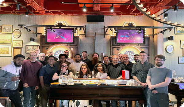 Group of coworkers smiling and posing together at a bar with dartboards and food in the background.