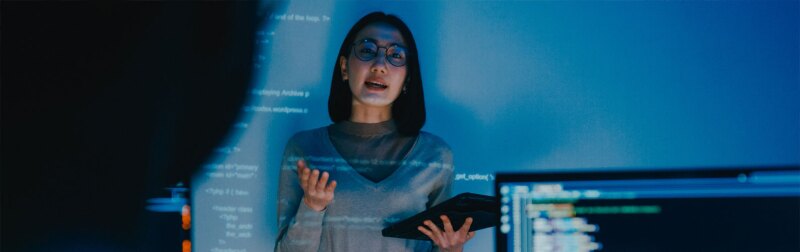 Woman speaking to others stands in front of a screen with code being projected onto it.