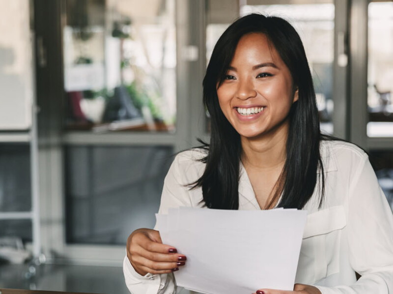 A young woman is smiling and holding a resume while sitting in front of directors during a corporate job interview.