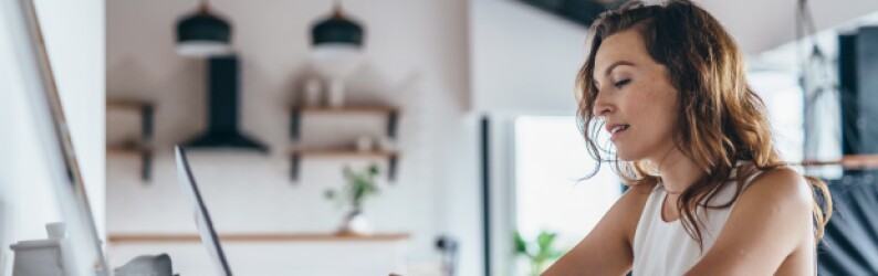 woman in white tank top seated at kitchen table completing coursework on laptop
