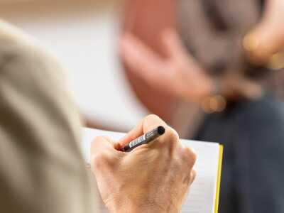 Looking closely over the shoulder of a counselor who takes notes while a woman sits across from them speaking.