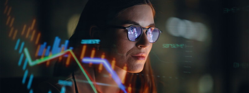 A woman in glasses reviews financial figures reflected on a transparent screen.
