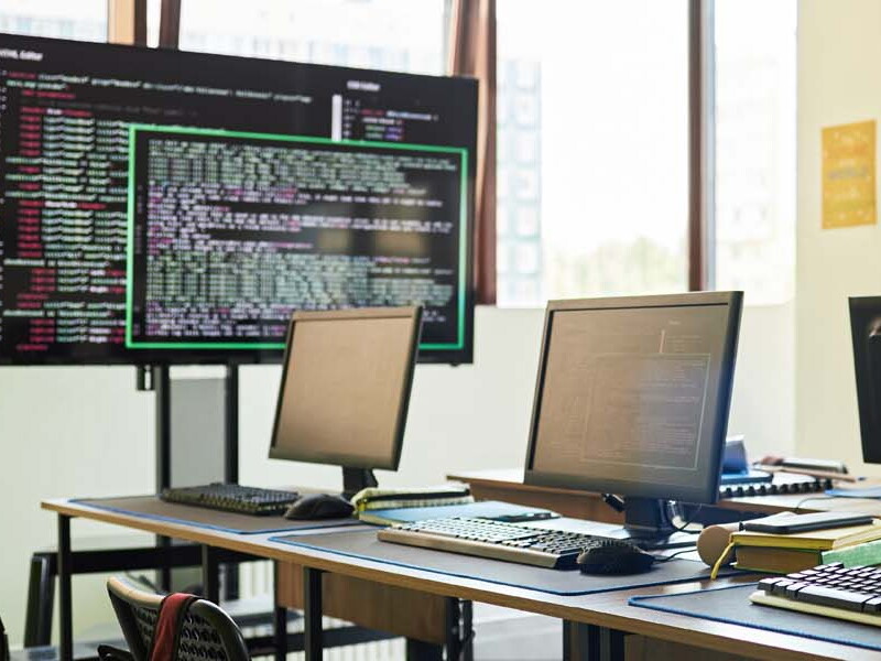 A sunny classroom is filled with various computers and screens displaying code.