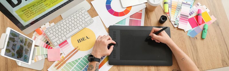 A designer working at a desk, using a drawing tablet and surrounded by various design tools and color swatches.