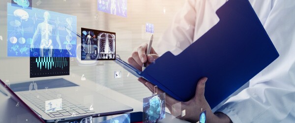 Person in lab coat with an open blue folder and pen in hand standing in front of a laptop