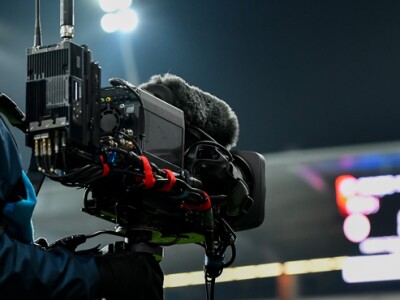 Man holding a camera at a sporting event, with a jumbotron in the distance