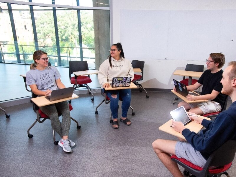 Students in desks seated in a circle and working on laptops and tablets