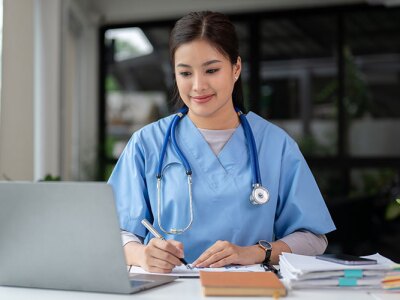 A nurse in scrubs is working on a laptop and taking notes.