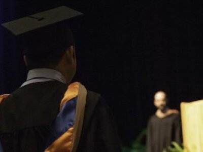 two-MBA-graduates-in-caps-and-gowns-with-backs-to-viewer-and-podium-and-graduation-speaker-in-foreground