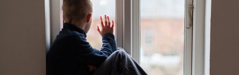 Young boy sitting on a windowsill looks out the window on a rainy day.