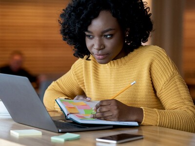 A woman in a cafe setting, focused on her laptop and notebook, searching for job opportunities