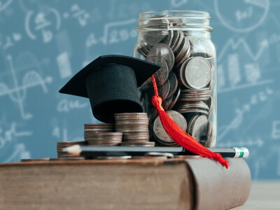 A tiny graduation cap, stacks of coins and a glass jar filled with coins are stacked up on a book.
