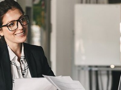 woman-in-glasses-and-black-blazer-smiling-at-woman-holding-papers-in-front-of-white-board