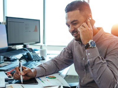 Male financial analyst discussing financial reports with colleague over mobile phone sitting at his office desk.