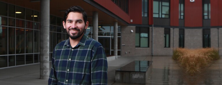 Man wearing plaid button down stands outside in front of a building