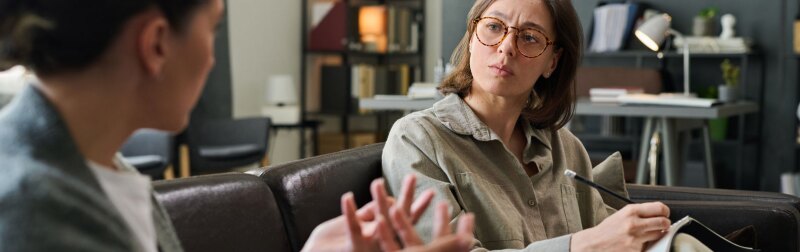 Two women sitting on couch discussing treatment plan during counseling session in cozy office.