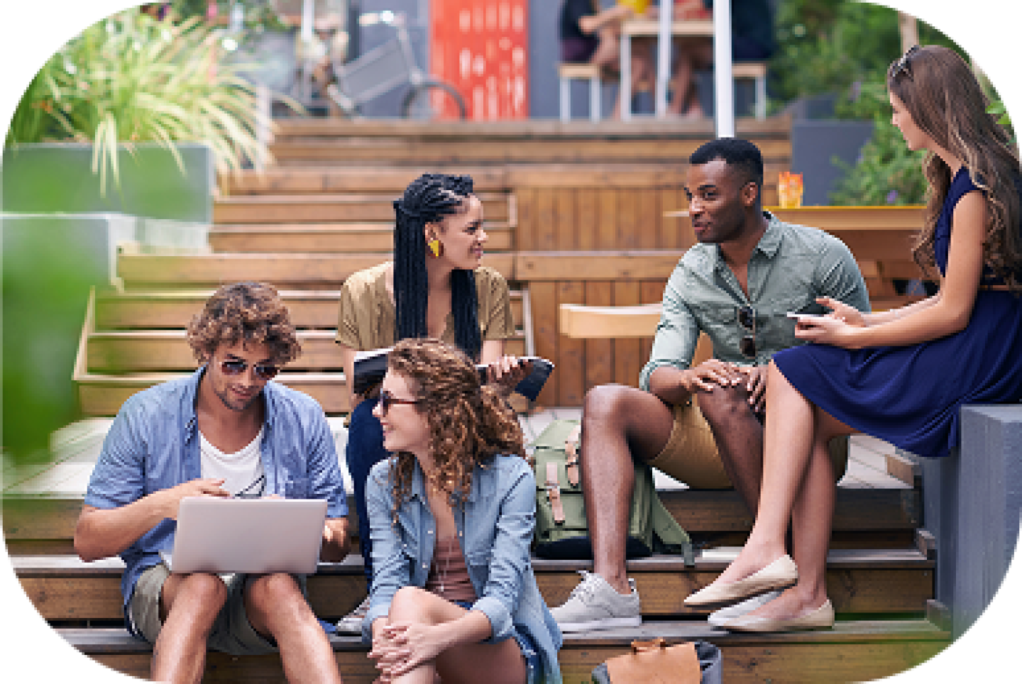 Group of college students sitting outdoors on campus steps, talking, studying, and using a laptop together.