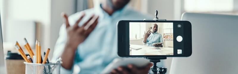 Man sitting at desk films himself with a cell phone camera.