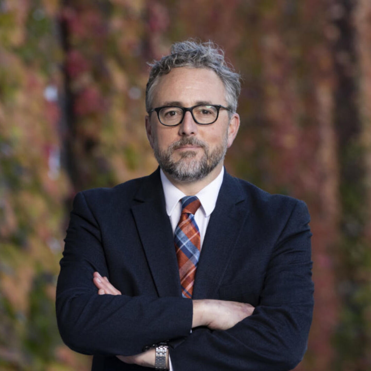 Professional headshot of a man with gray hair and glasses in a navy suit and plaid tie, standing outdoors with autumn foliage background.