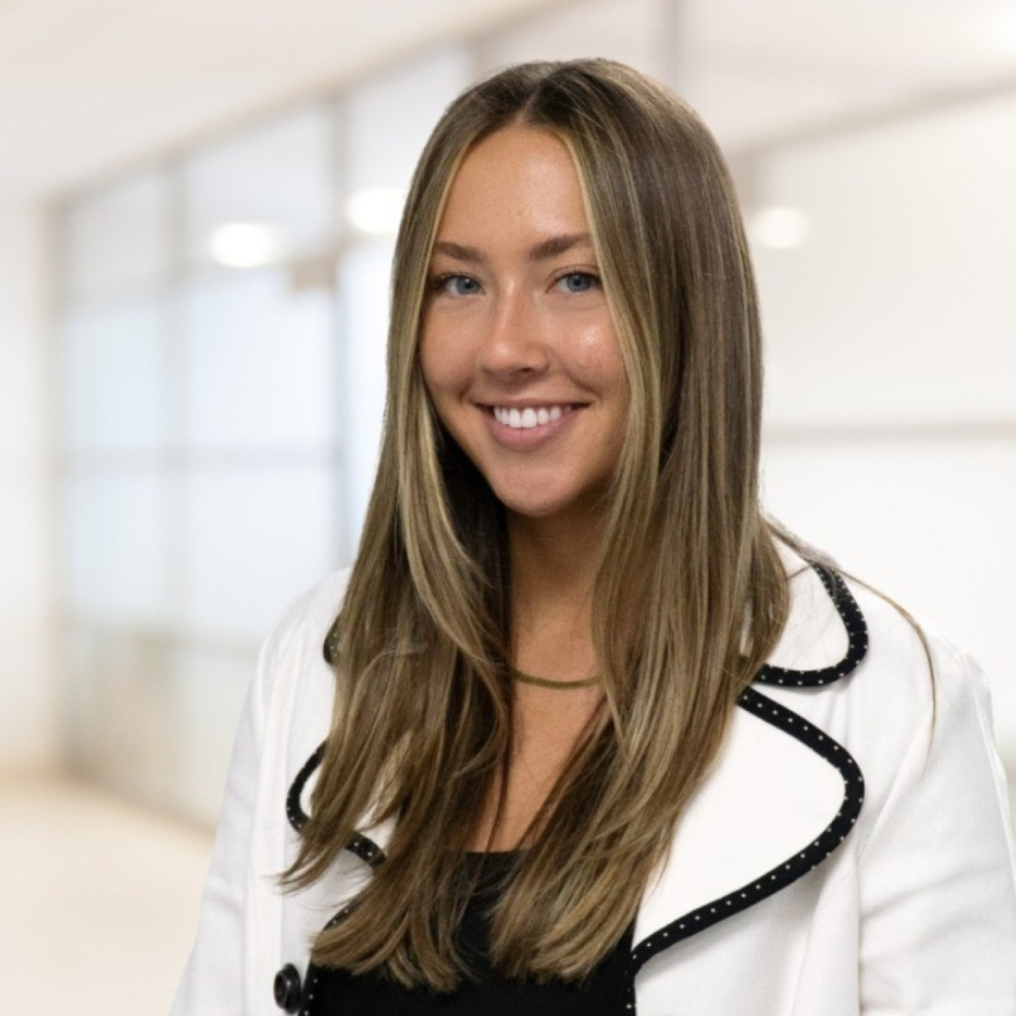 Headshot of Alexa Bauman, with long brown hair, wearing a light blazer over a black top, in a bright, modern office setting.