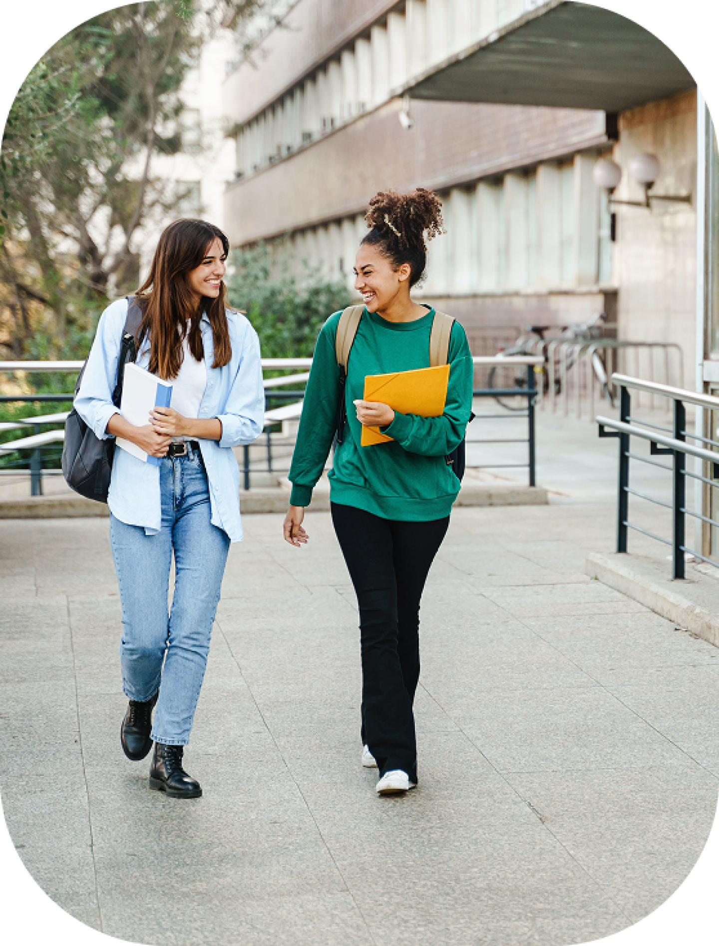 Two college students walking outdoors on campus, smiling and carrying notebooks and backpacks between classes.