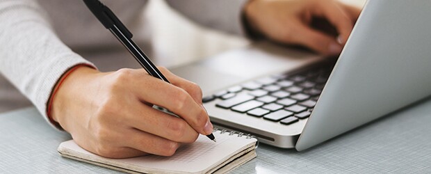 A person's left hand types on a laptop while the right hand jots notes in a notebook