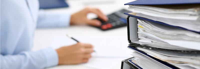Binders with papers are waiting in the foreground, woman conducting an audit in the background