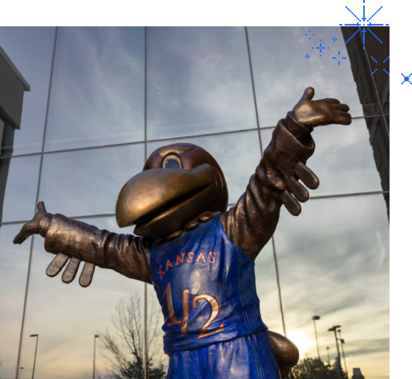 Jayhawk statue with arms raised in front of building reflecting the sky
