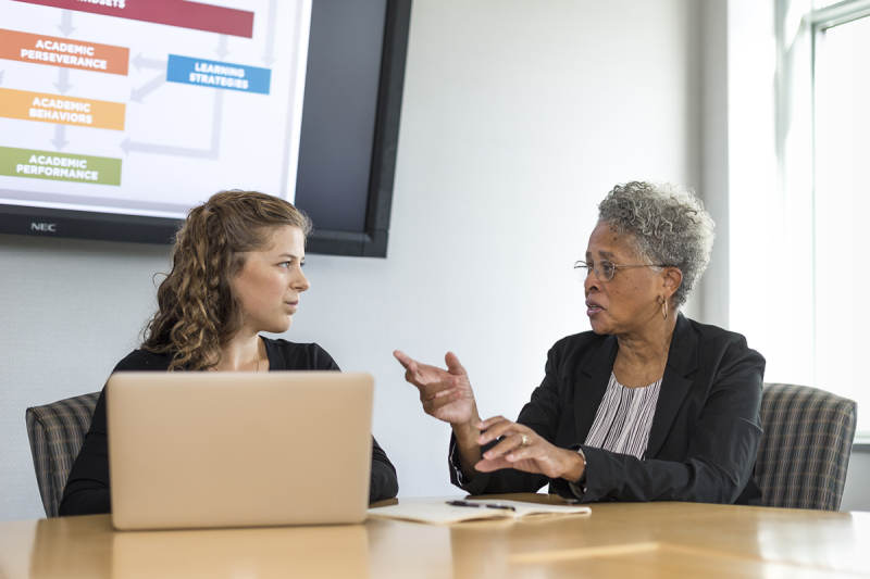 Two woman collaborating at conference table in front of video display of flowchart