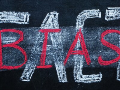 Erased word 'FACT' on a chalkboard, partially overwritten by the word 'BIAS' in red chalk