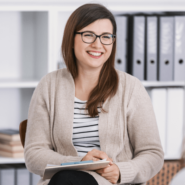 Woman in cardigan and glasses smiling while holding notepad
