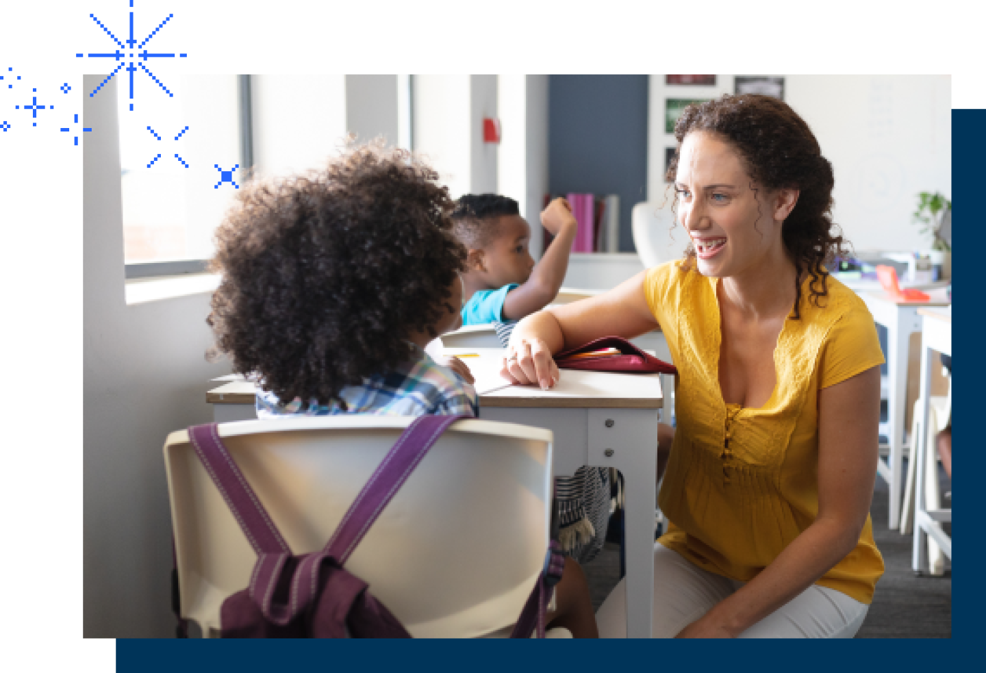A female teacher kneels next to a female student's desk.