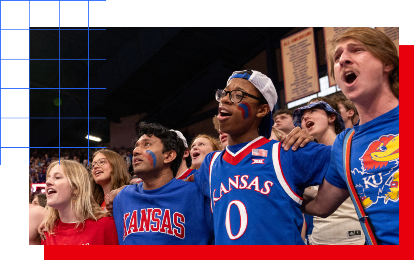 Group of Kansas Jayhawks fans standing arm-in-arm in a crowded arena, singing or cheering, many wearing blue KU shirts and face paint.