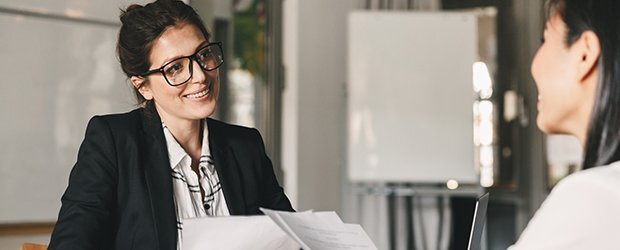 woman-in-glasses-and-black-blazer-smiling-at-woman-holding-papers-in-front-of-white-board