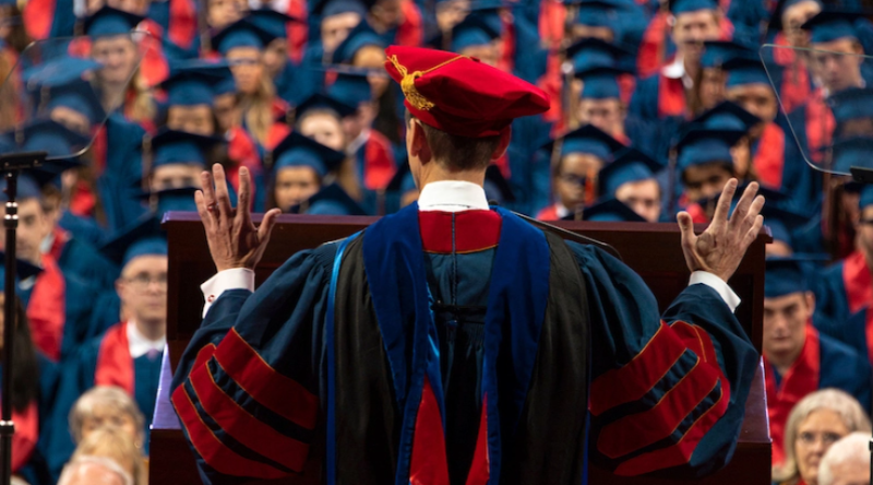 Distinguished high-ranking faculty adorned in full graduation regalia, delivering a commencement speech to the graduating class.