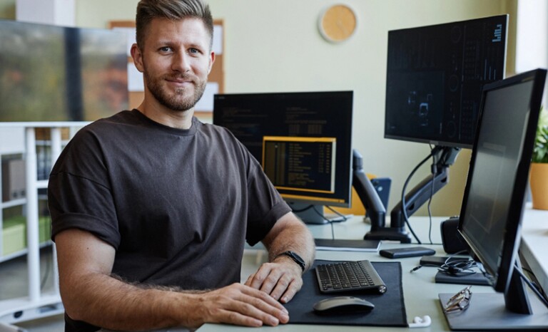 Man in T-shirt seated at desk with multiple computer monitors and keyboard