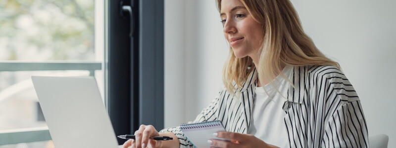 Woman working on a laptop with a notepad in her hand