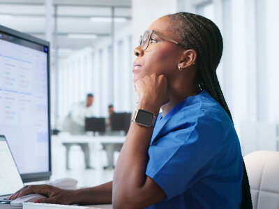 A woman in blue scrubs sits in front of a laptop and two external computer screens