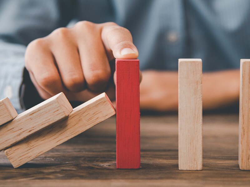 A man sitting at the table has his finger on top of the red block, stopping the remaining blocks from falling over.