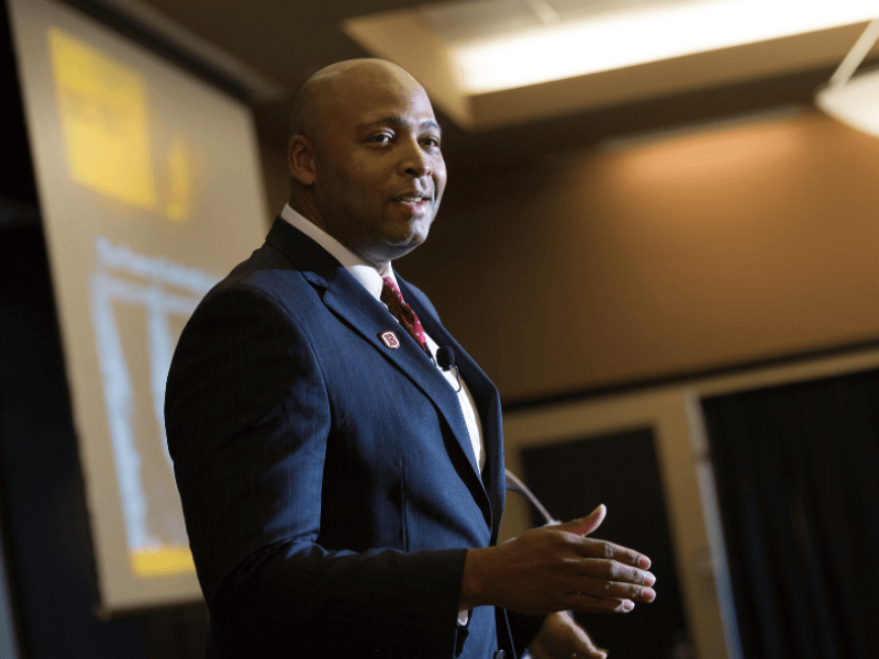 Man in blue suit with Bradley University pin delivering speech in front of screen