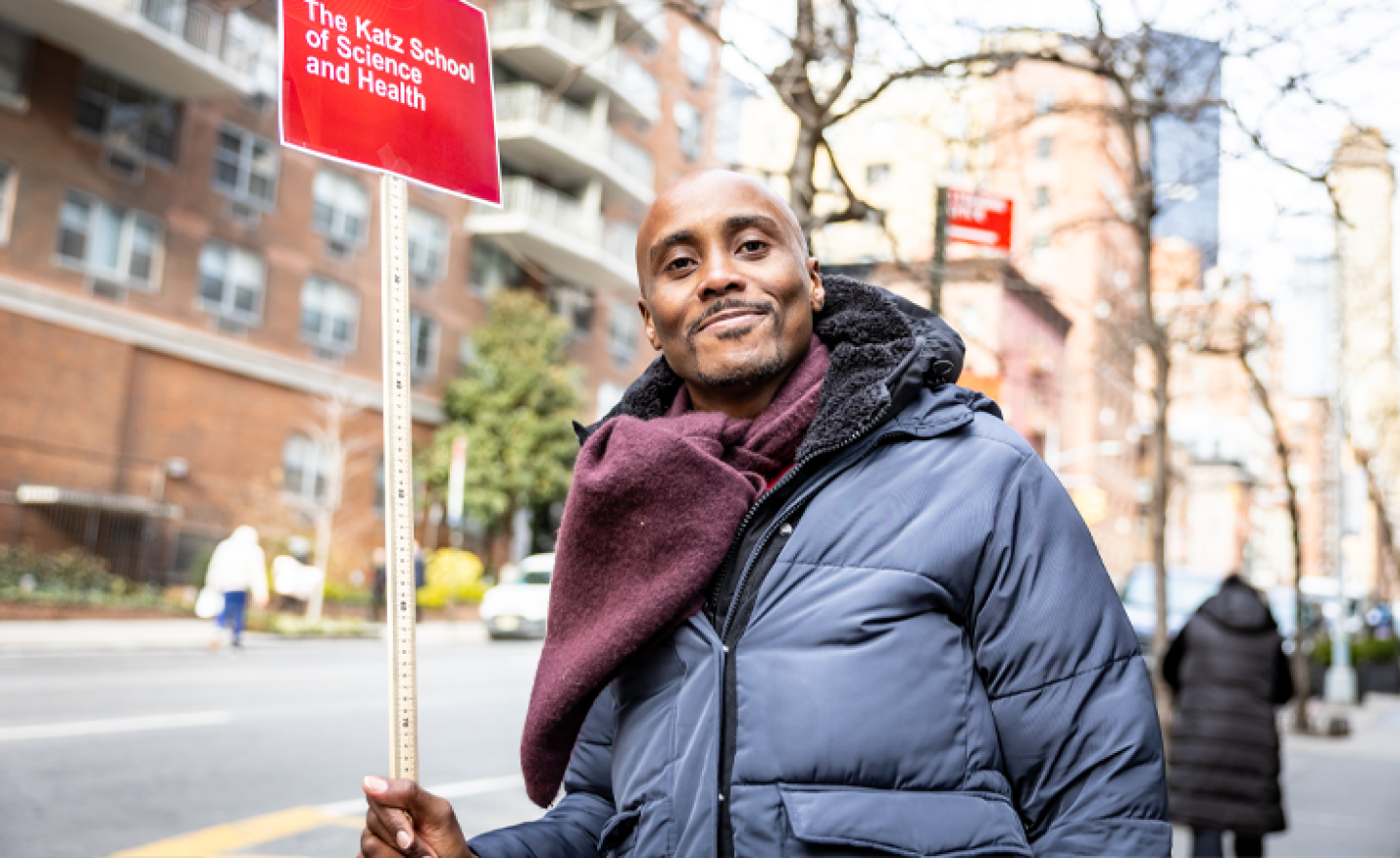 A male Yeshiva University student standing on the street while holding a sign that reads The Katz School of Science and Health.